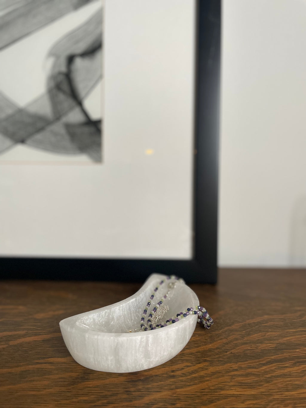 White Selenite crescent moon shaped dish with a necklace on a wooden surface, with a blurred abstract painting in the background.