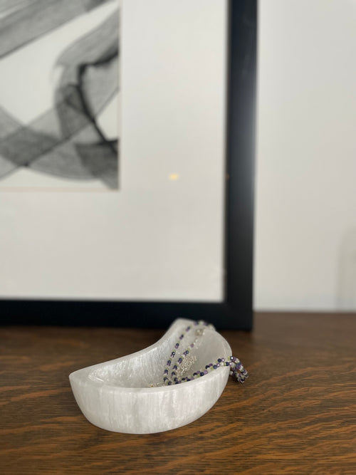 White Selenite crescent moon shaped dish with a necklace on a wooden surface, with a blurred abstract painting in the background.